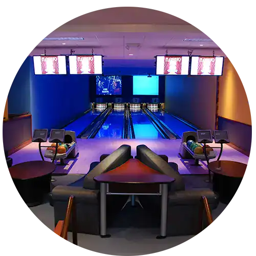 Four bowling lanes lit with blue and purple light. Bowling balls sit on the return machines and TVs hang in front and projected screens hang over the back of the lanes.